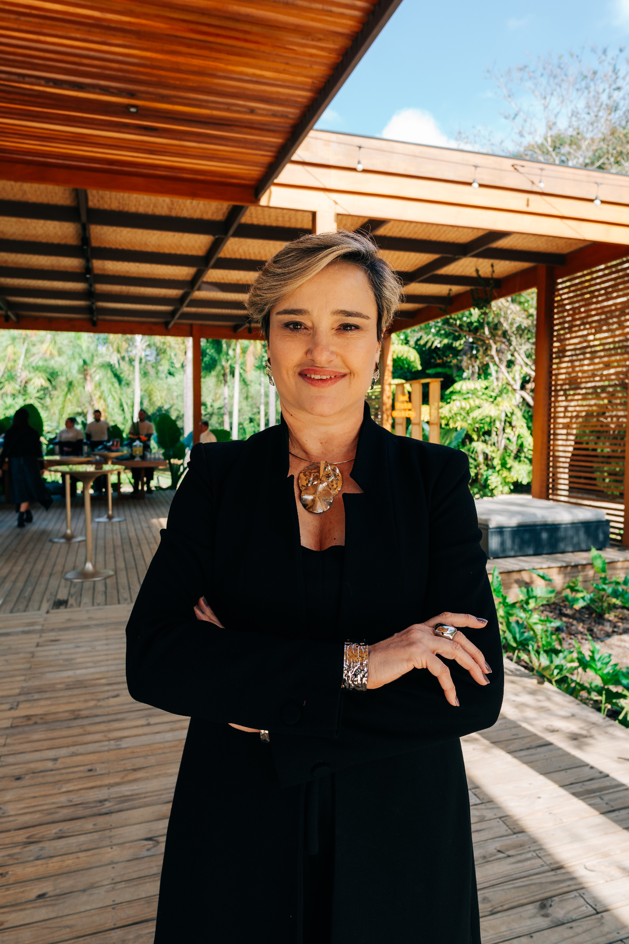 Mulher madura sorridente, braços cruzados, em ambiente natural com madeira e jardim